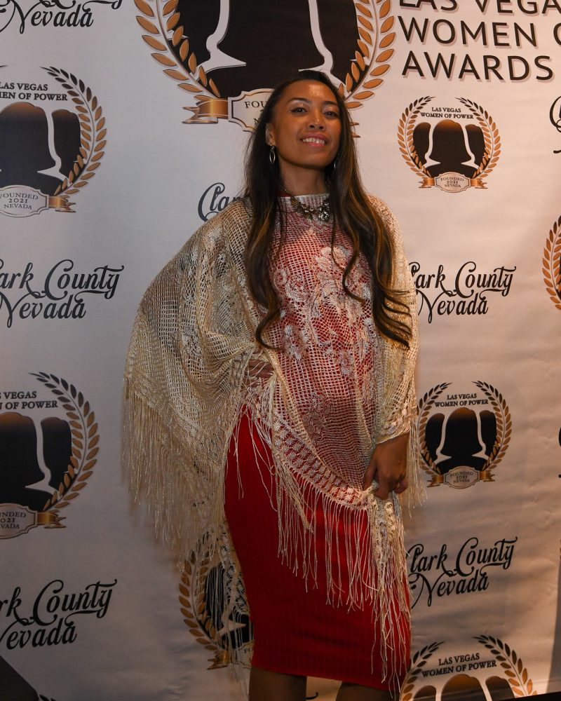 Woman in a fringed shawl and red dress posing at the Las Vegas Women of Power Awards event backdrop.